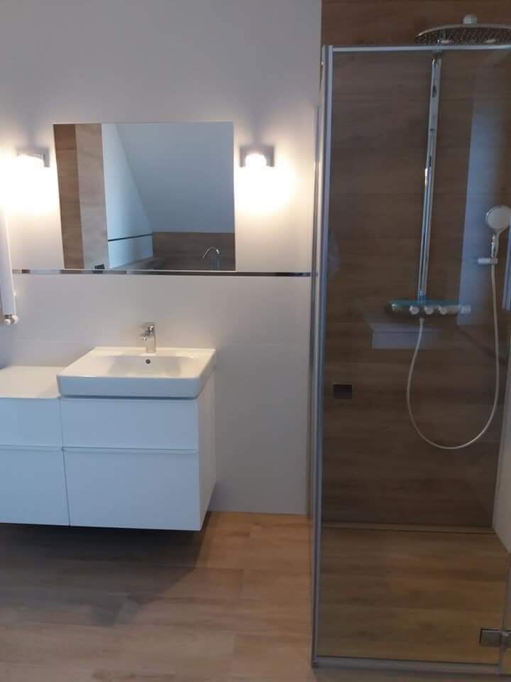 Nowoczesna łazienka z kabiną prysznicową typu walk-in, białą szafką pod umywalkę i lustrem z oświetleniem punktowym, wykończona płytkami imitującymi jasne drewno.
