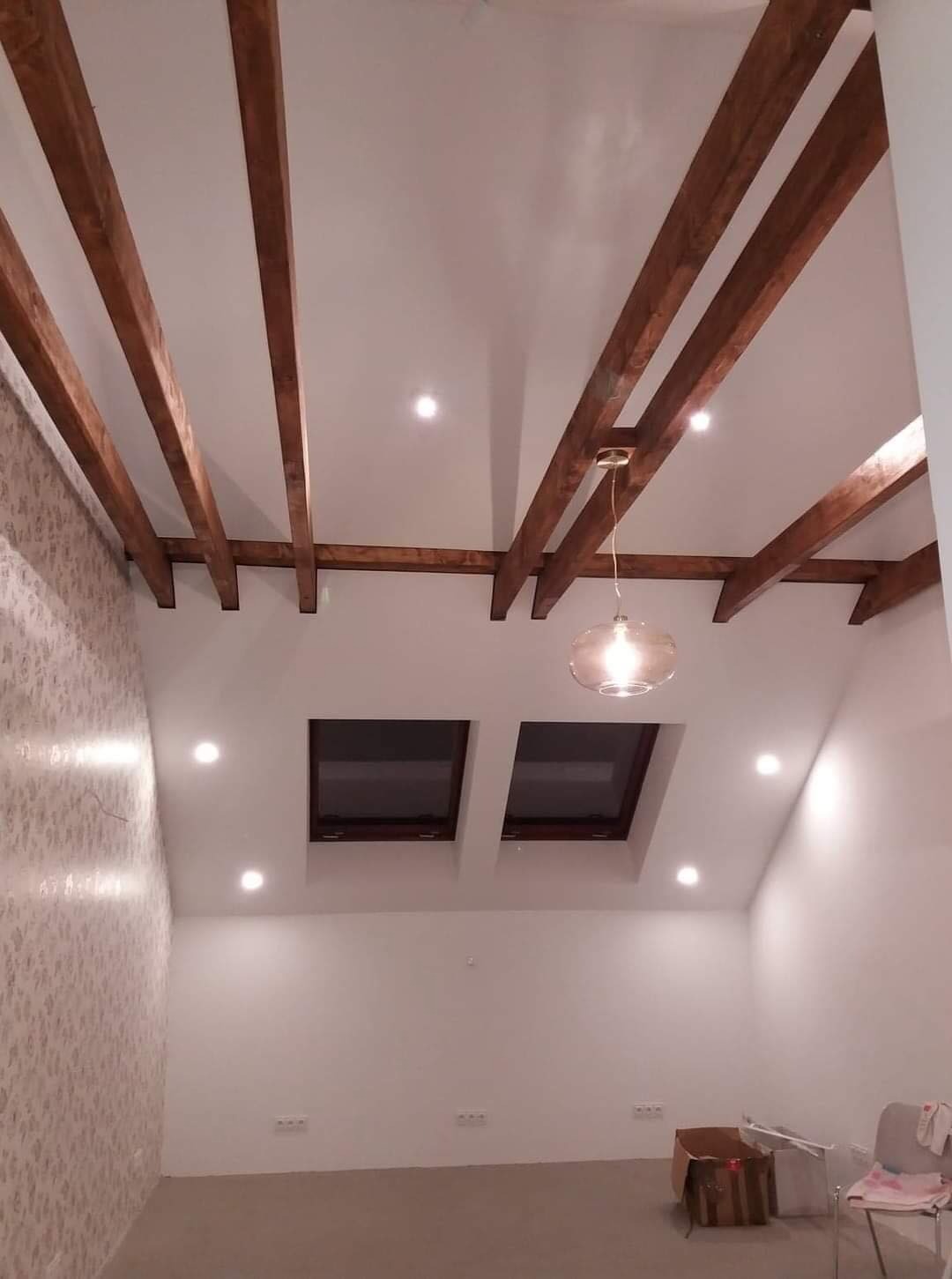 Pokój na poddaszu po remoncie z drewnianymi belkami na suficie, dwoma oknami dachowymi i tapetą w delikatny wzór na jednej ze ścian.