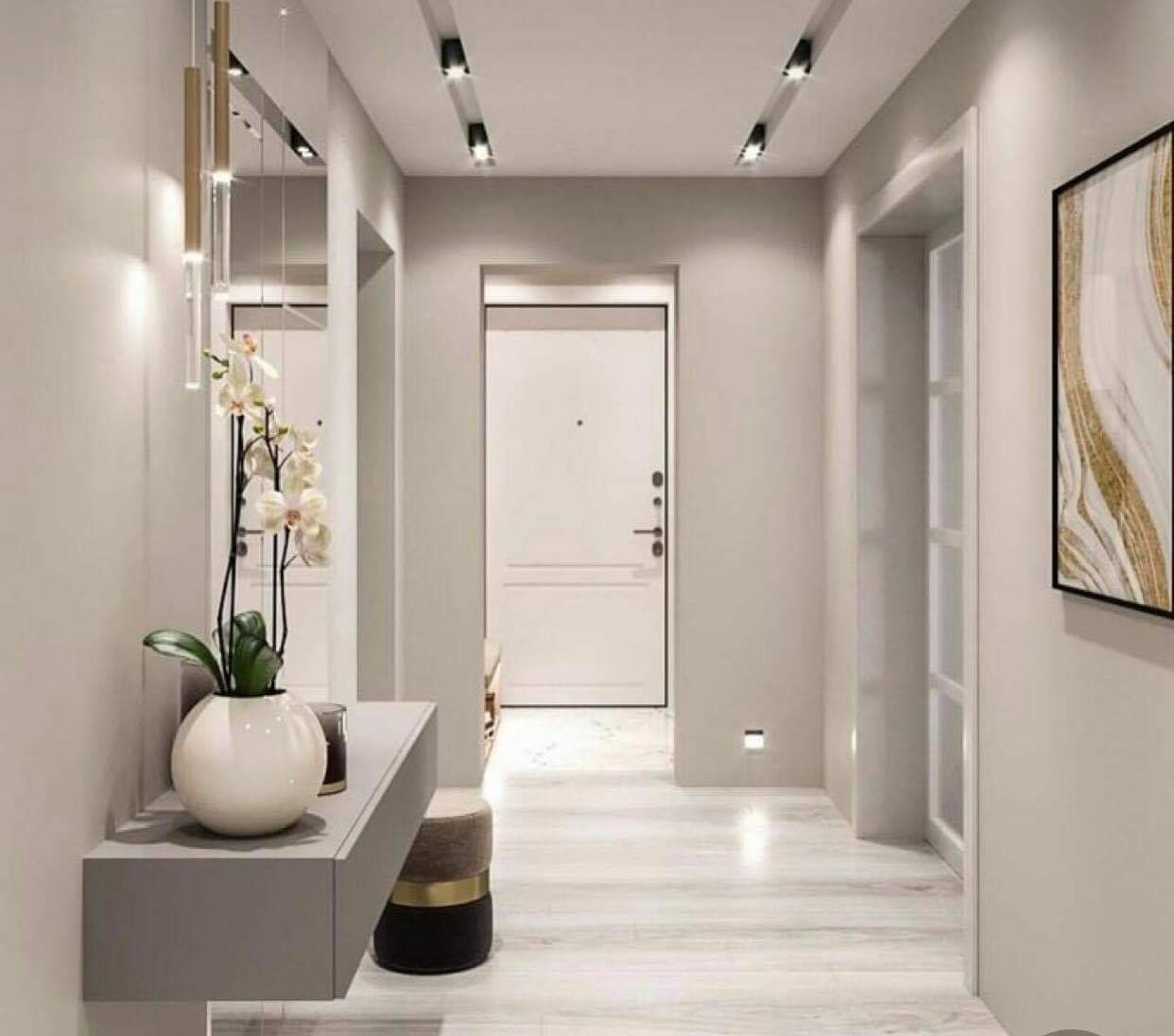 Nowoczesny przedpokój w jasnych odcieniach szarości z białymi drzwiami, marmurową podłogą, minimalistyczną konsolą z kwiatami i złotymi akcentami w oświetleniu oraz dekoracjach.