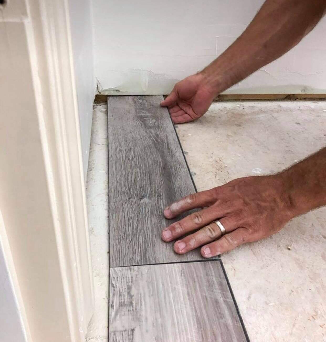 Układanie paneli podłogowych w odcieniach szarości przy białej ścianie, widoczne dłonie z obrączką i niedokończona podłoga.