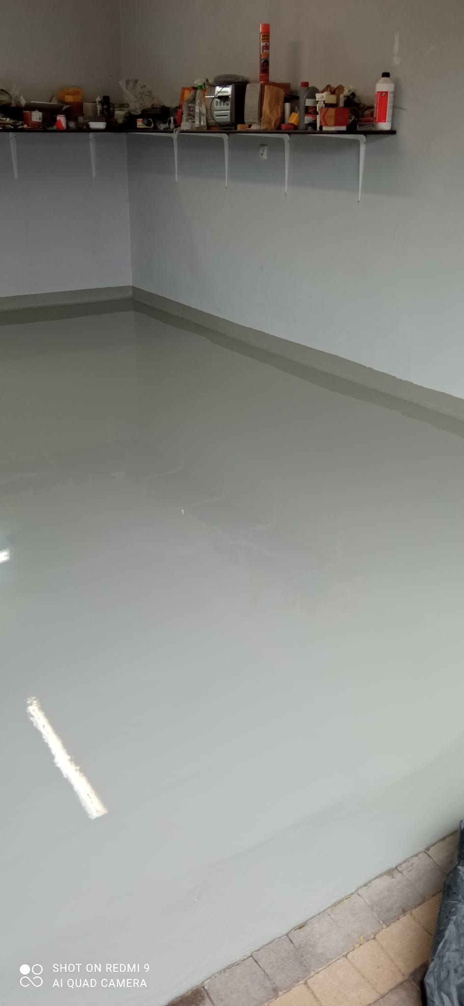 Świeżo wylana, jasnoszara posadzka żywiczna w pomieszczeniu z półką na ścianie, widoczne refleksy światła na gładkiej powierzchni.