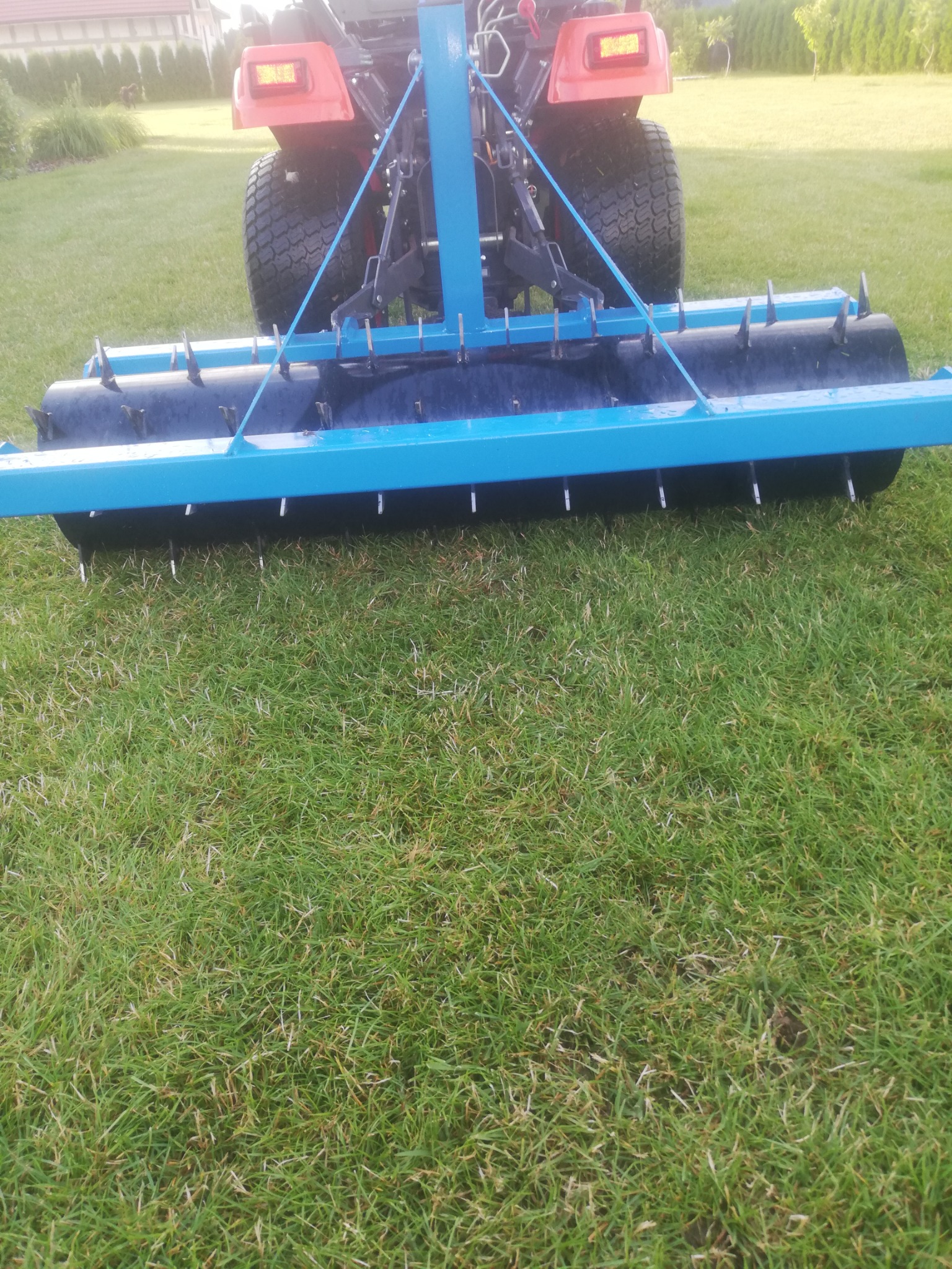 Czerwony traktor z niebieskim wałem kolczastym na trawniku, widok z tyłu, w tle dom jednorodzinny.