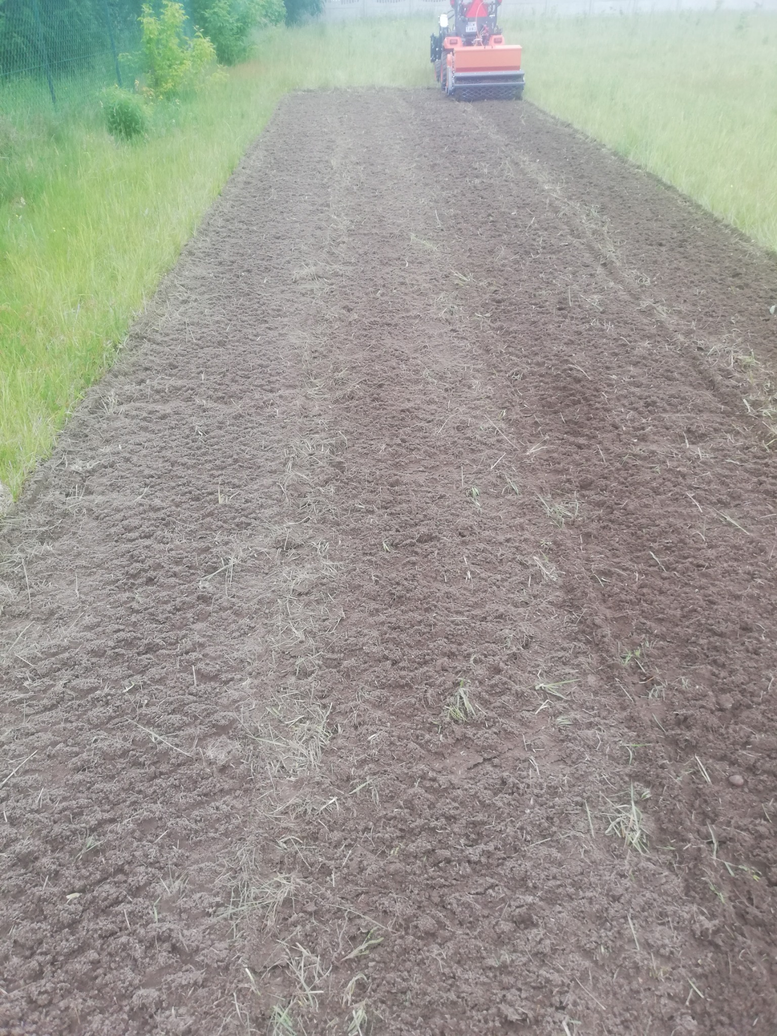 Przygotowana gleba pod trawnik z rolki, widoczny mały traktor z glebogryzarką w oddali, świeżo przekopana ziemia z resztkami trawy.