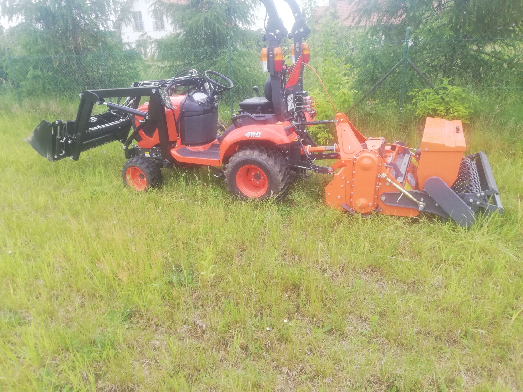 Pomarańczowy traktor 4WD z zamontowanym z przodu ładowaczem i kosiarką rotacyjną na tle wysokiej trawy i drzew