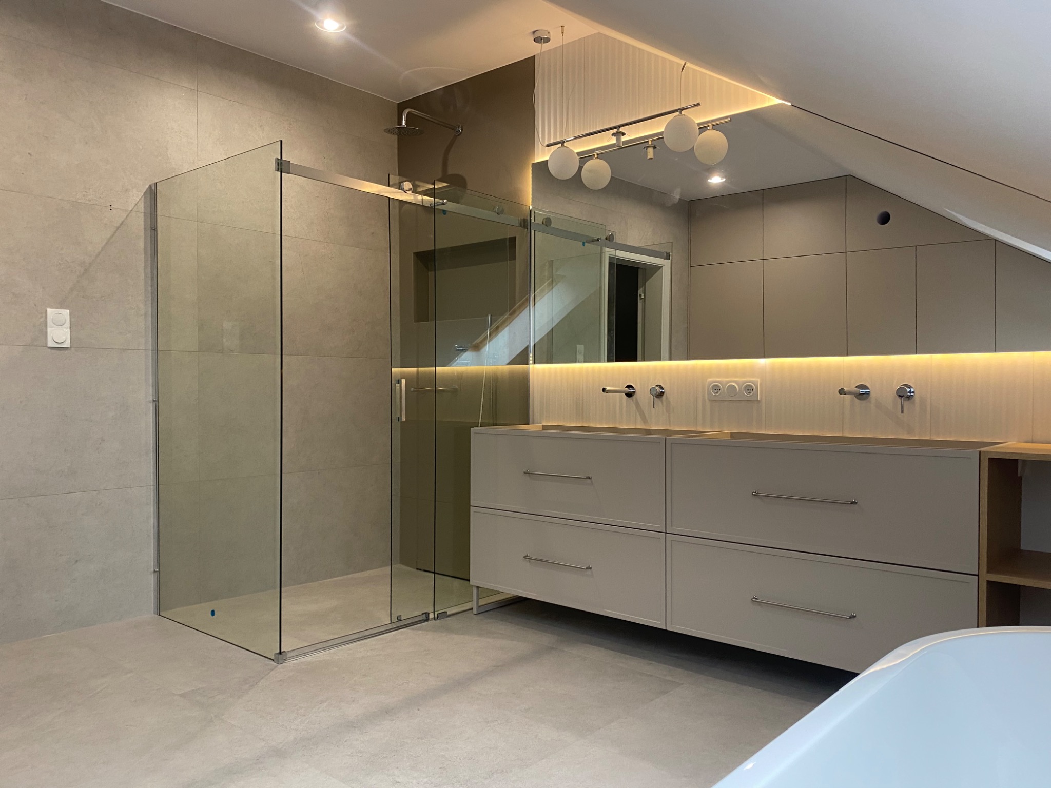 Nowoczesna łazienka na poddaszu z kabiną prysznicową ze szklanymi przesuwnymi drzwiami, podwójną umywalką z szafką w odcieniach szarości i oświetleniem LED pod lustrem.