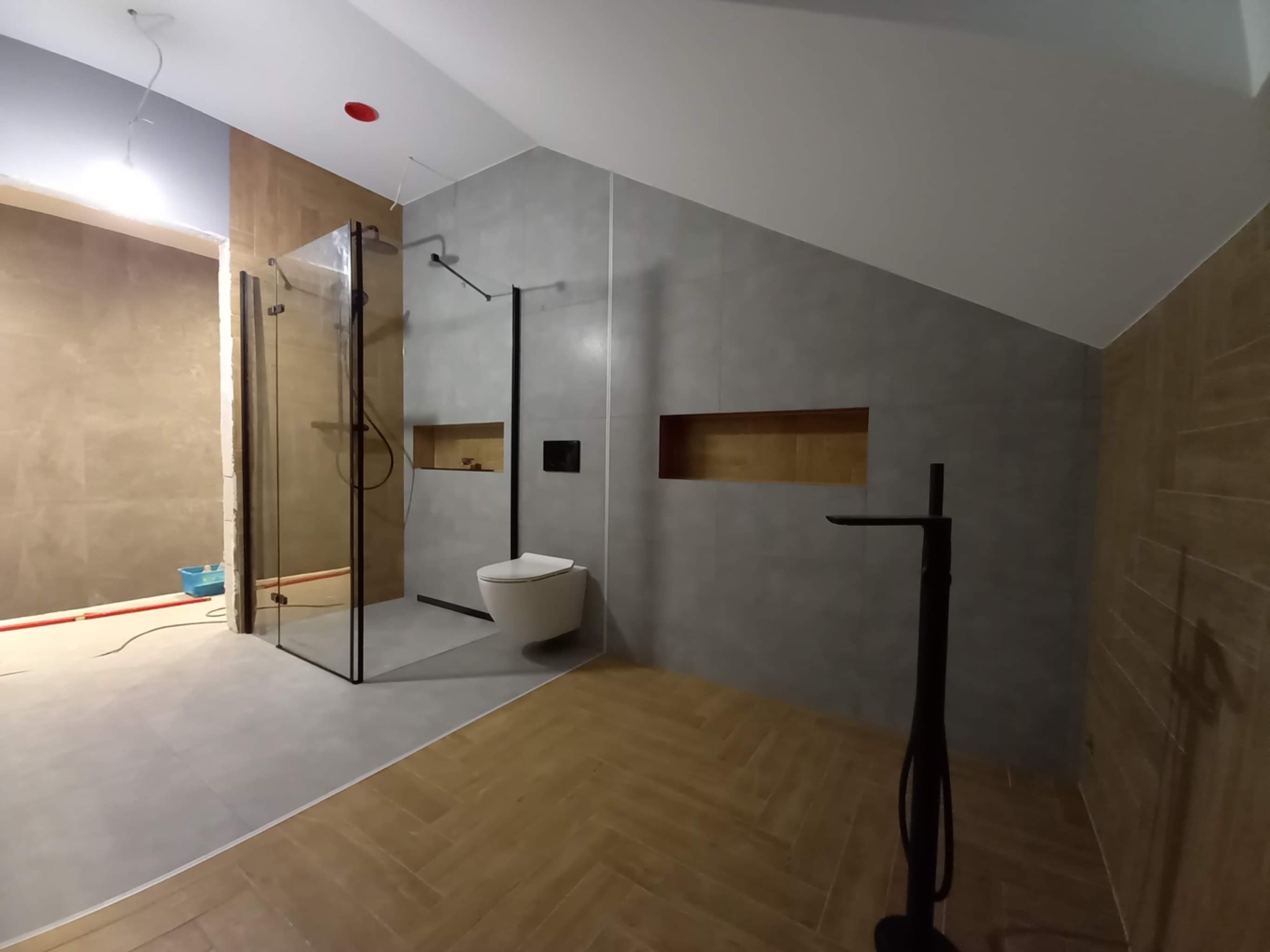 Nowoczesna łazienka w trakcie wykańczania: szklana kabina prysznicowa z czarnymi profilami, podwieszana toaleta, drewnopodobna podłoga ułożona w jodełkę, szare płytki ścienne, widoczne...