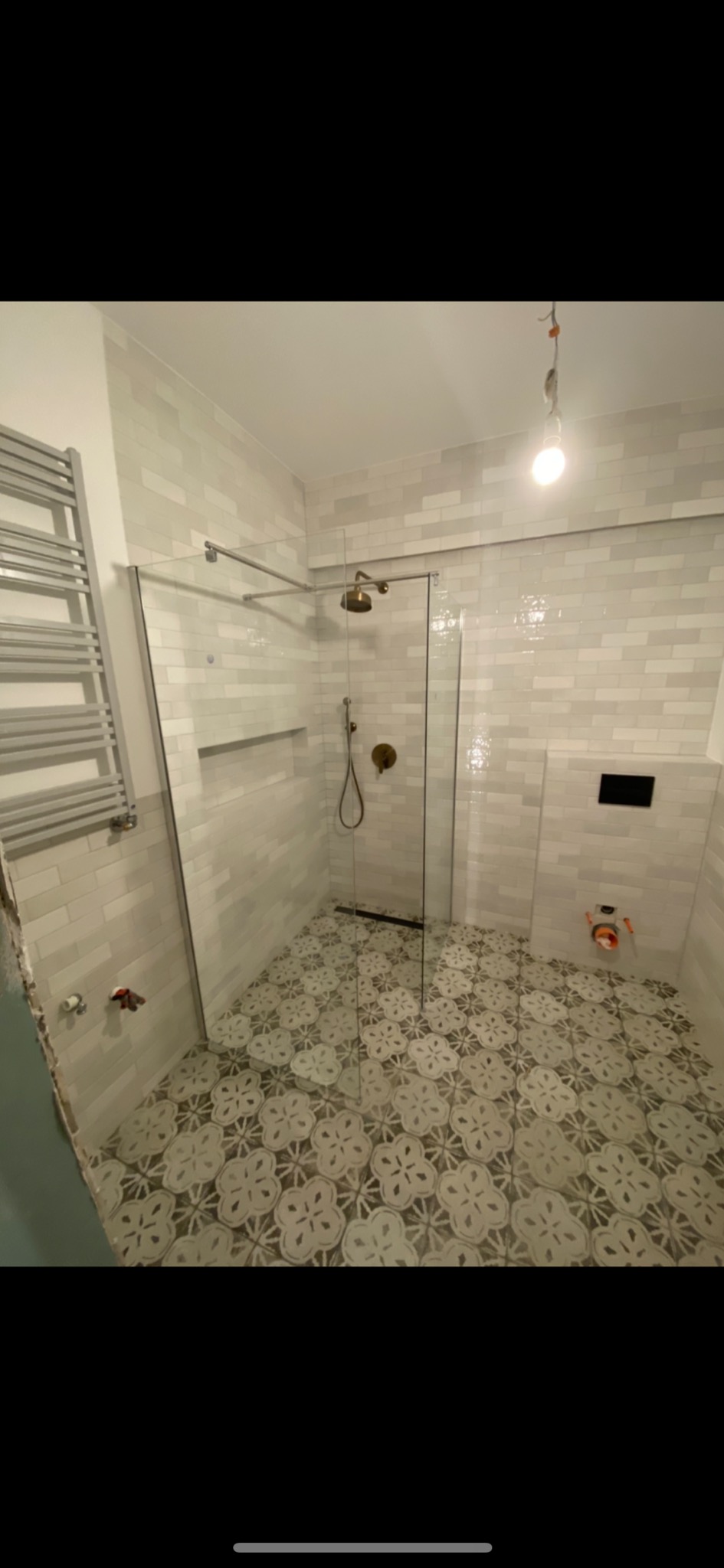 Ukończona łazienka z kabiną prysznicową ze szklaną ścianką, jasnymi płytkami na ścianach i wzorzystą podłogą w kwiatowy deseń. Widoczny grzejnik drabinkowy, złota armatura i wnęka w ścianie.