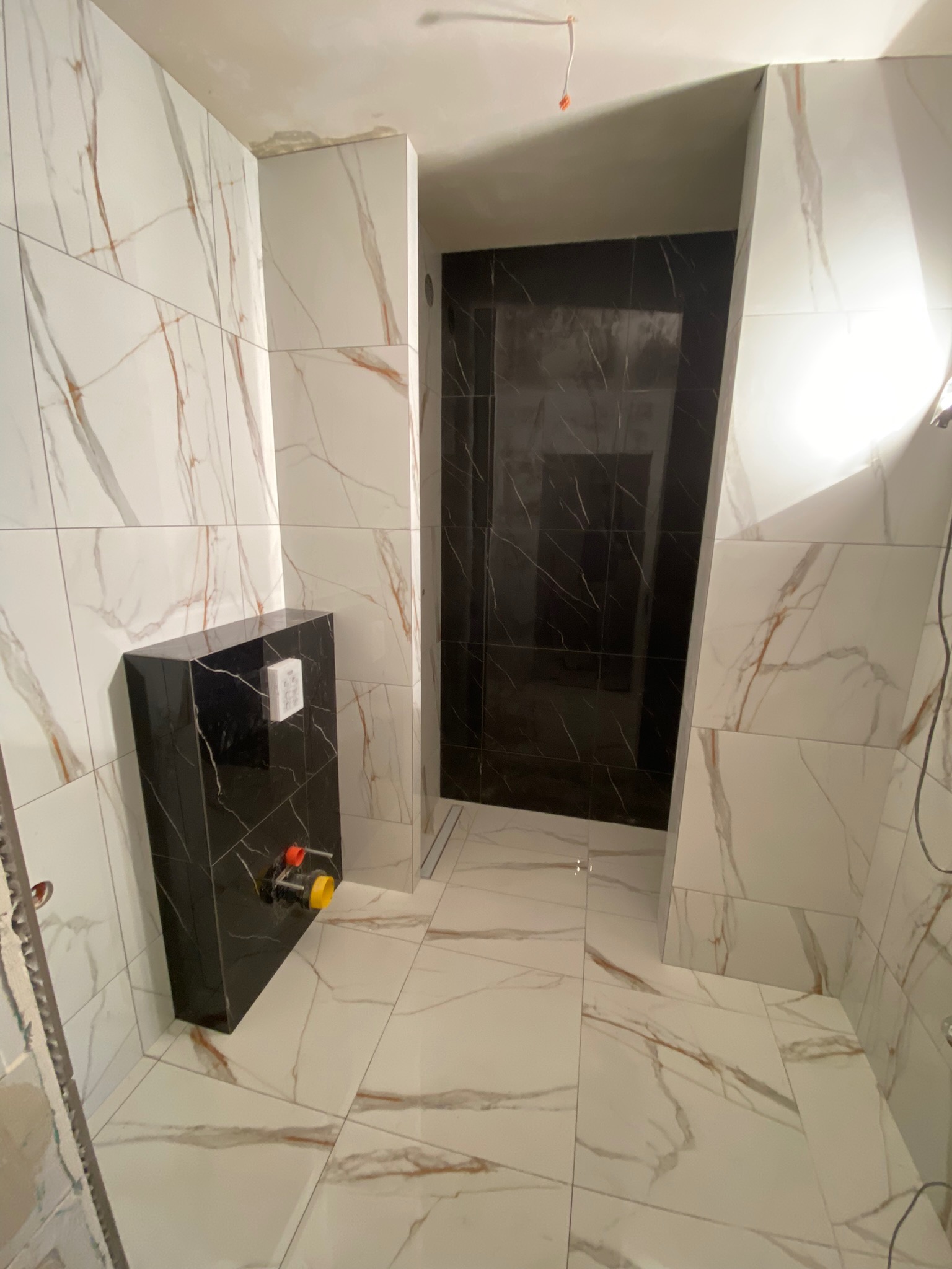 Częściowo wykończona łazienka z białymi płytkami imitującymi marmur z brązowymi żyłami na ścianach i podłodze, czarna obudowa stelaża podtynkowego WC, widoczny odpływ liniowy w kabinie prysznicowej...