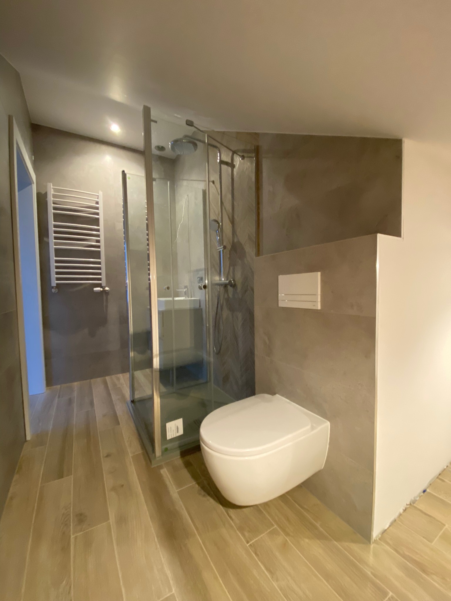 Nowoczesna łazienka z kabiną prysznicową typu walk-in, wiszącą toaletą i grzejnikiem drabinkowym, wykończona płytkami imitującymi drewno na podłodze i szarymi płytkami na ścianach, oświetlona...