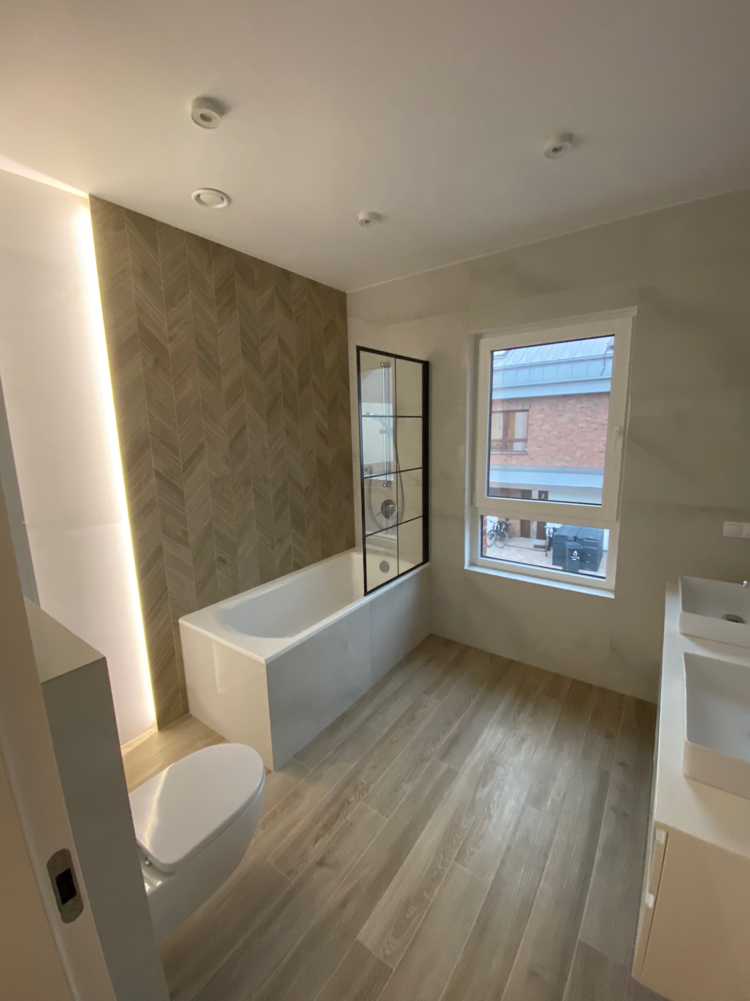 Nowoczesna łazienka z wanną z czarną ramą kabiny, umywalką nablatową i podświetloną ścianą z wzorem jodełki, drewnopodobna podłoga z płytek.