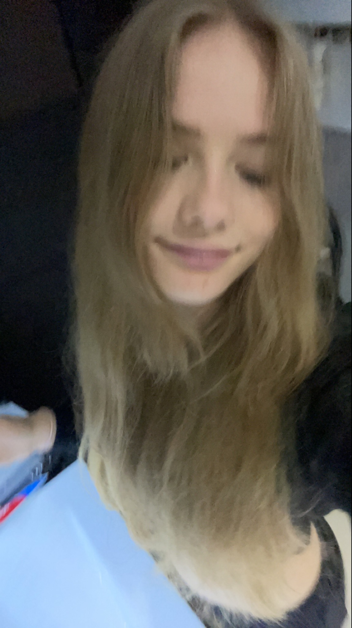 Portret młodej kobiety z długimi blond włosami, uśmiechająca się delikatnie w kierunku obiektywu, z fragmentem białego pojemnika widocznym w dolnej części kadru.