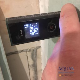 Aqua Construction - Pomiar odległości laserowym miernikiem Zamo na tle surowej ściany w trakcie prac wykończeniowych, wyświetlający 1.698 metra.