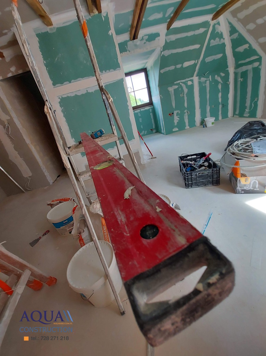 Poddasze w trakcie prac wykończeniowych: ściany pokryte płytami gipsowo-kartonowymi, częściowo zaszpachlowane, widoczna drabina, poziomica, wiadra z zaprawą i narzędzia.