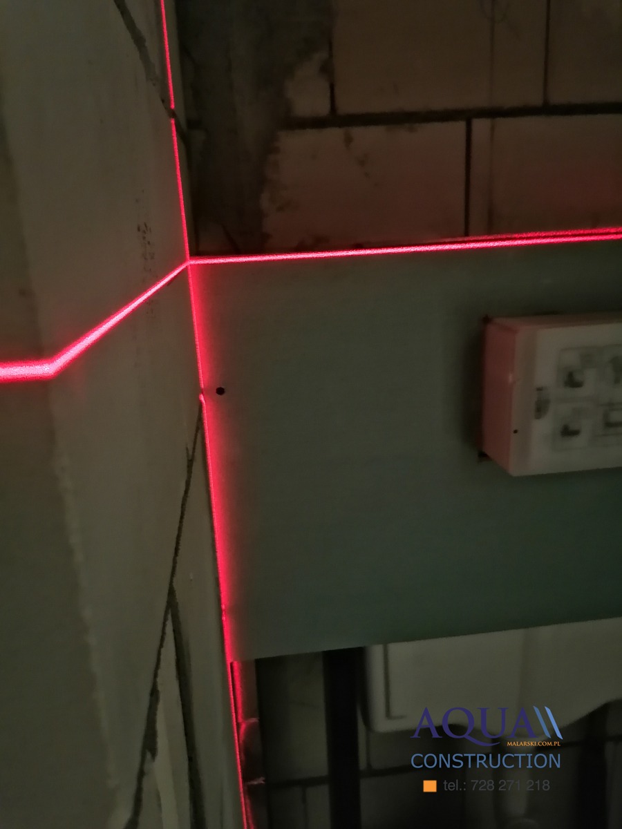 Czerwone linie lasera wyznaczają poziom na ścianie w trakcie prac remontowych, widoczny fragment ściany z bloczków i logo firmy w rogu.
