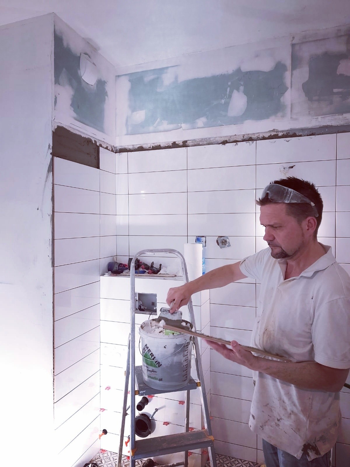 Mężczyzna w okularach ochronnych nabiera zaprawę z wiadra stojącego na drabinie, w tle częściowo wyłożona białymi płytkami ściana łazienki i przygotowane do położenia fragmenty ścian.