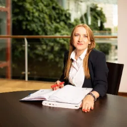 Kobieta z długimi rudymi włosami siedzi przy ciemnym stole w nowoczesnym biurze, trzyma czerwony długopis nad otwartym kalendarzem leżącym na białym laptopie.