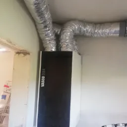 Centrala wentylacyjna Vasco DX4 z podłączonymi aluminiowymi przewodami wentylacyjnymi, widoczne instalacje wodne w tle, surowy stan wykończenia wnętrza.