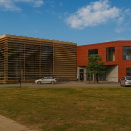 D.S.A Szymon Dądela - Nowoczesny budynek biurowy z elewacją w odcieniach brązu i pomarańczy, otoczony zielonym trawnikiem, z zaparkowanymi samochodami i rowerami. Architektura minimalistyczna.