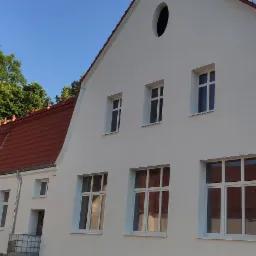 Biały budynek z nową, czerwoną dachówką, widoczne obróbki blacharskie i okna. Na dachu widoczne okno dachowe. Część budynku jest otoczona rusztowaniem. W tle drzewa i błękitne niebo.