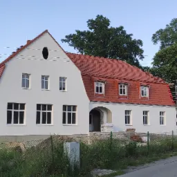 Odnowiony budynek z czerwoną dachówką, białą elewacją i owalnym oknem na poddaszu, częściowo widoczne rusztowanie przy dachu.