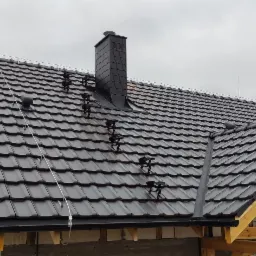 Nowy dach z czarnej dachówki ceramicznej z kominem i systemem odgromowym, widoczne drewniane elementy konstrukcji dachu w surowym stanie.