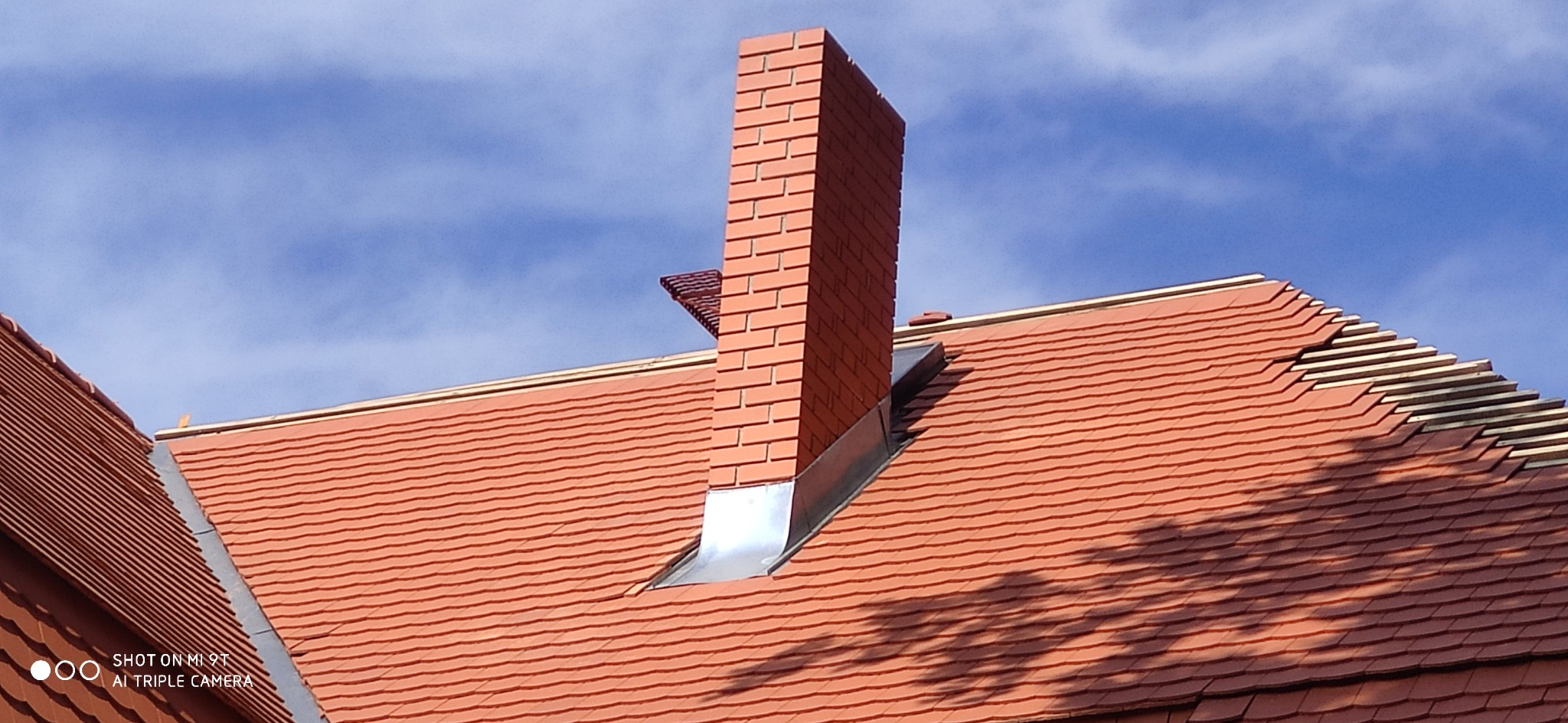 Ceglany komin z obróbką blacharską na dachu krytym czerwoną dachówką karpiówką, widoczne fragmenty łat i kontrłat, błękitne niebo w tle.