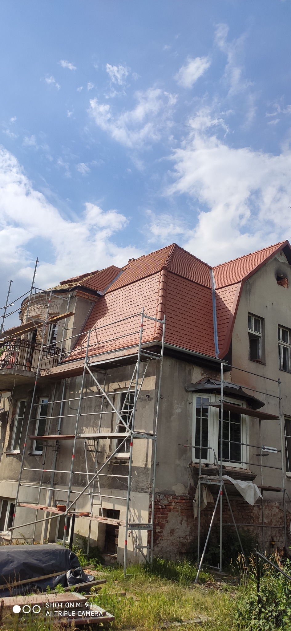 Dom w trakcie renowacji, z rusztowaniami, nową czerwoną dachówką na części dachu i szarą elewacją przed ukończeniem prac.