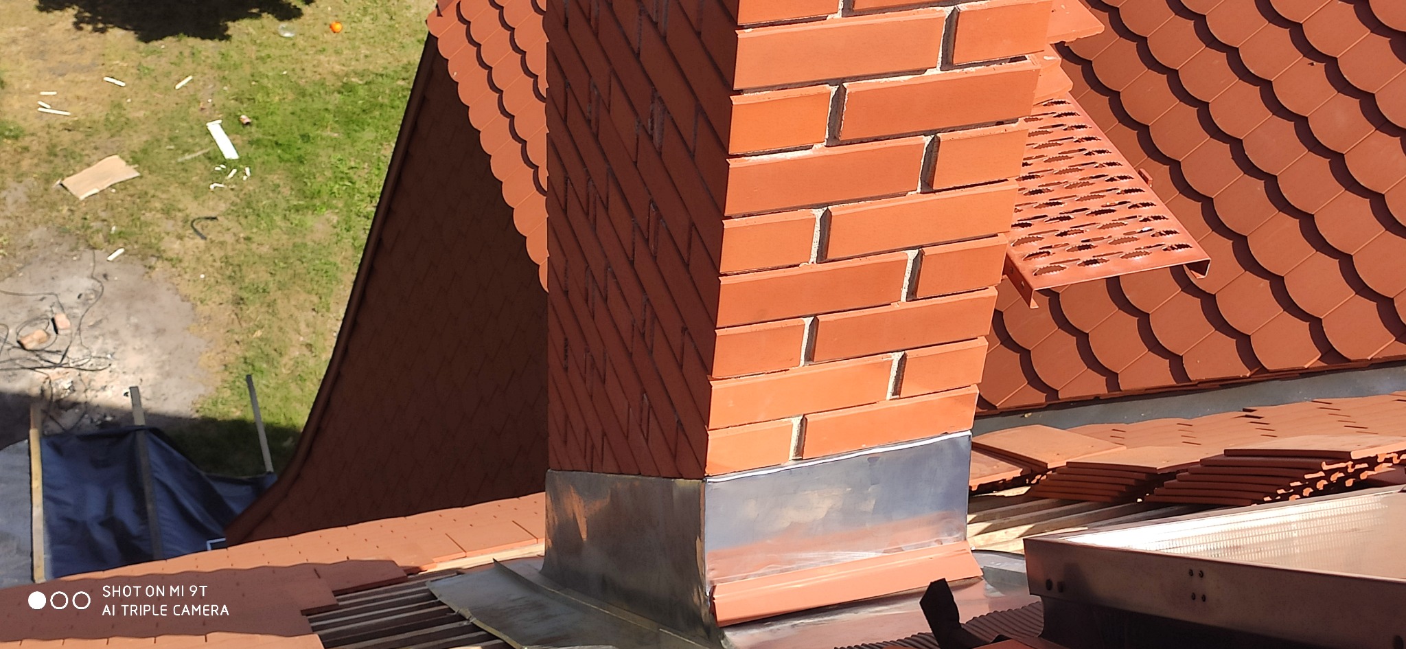 Widok z góry na komin z cegły klinkierowej z metalową obróbką u podstawy, otoczony nową, czerwoną dachówką karpiówką. W tle widoczny fragment trawnika z porozrzucanymi elementami budowlanymi.