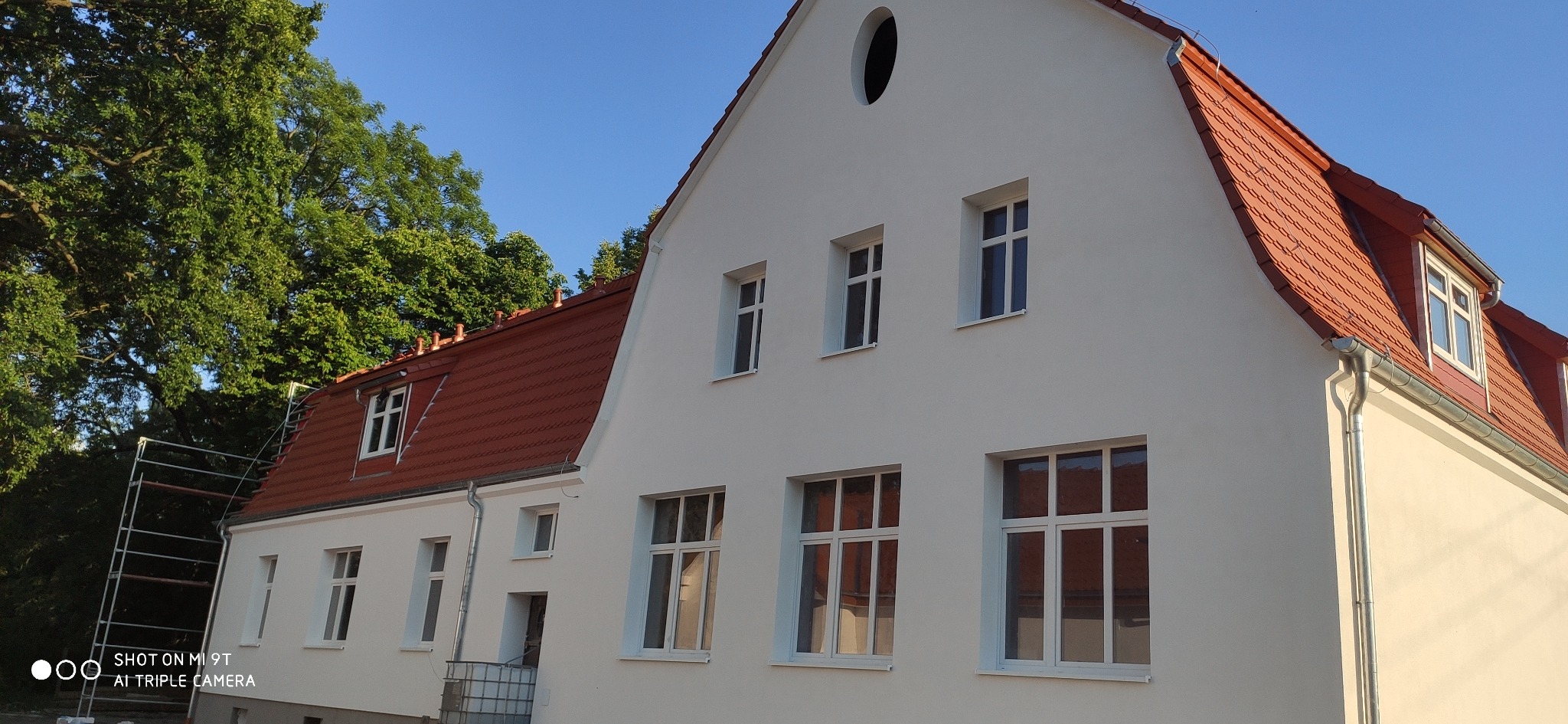 Biały budynek z nową, czerwoną dachówką, widoczne obróbki blacharskie i okna. Na dachu widoczne okno dachowe. Część budynku jest otoczona rusztowaniem. W tle drzewa i błękitne niebo.