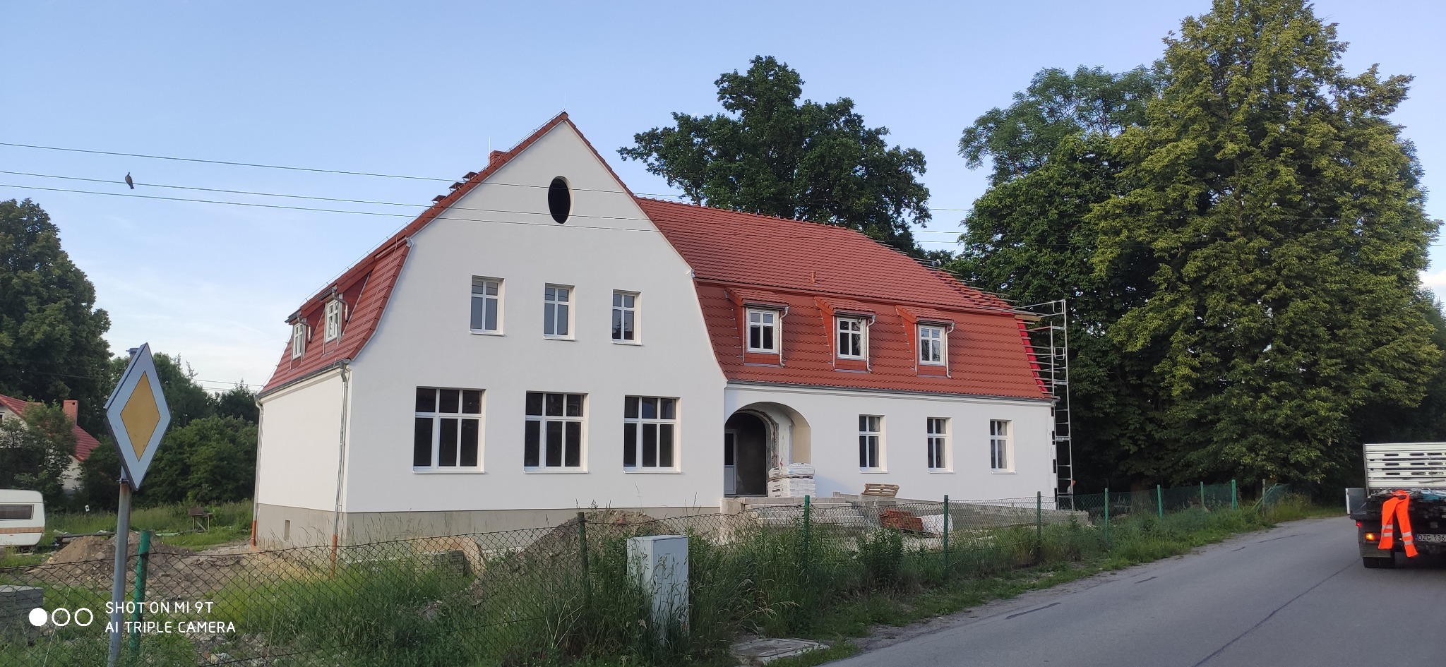 Odnowiony budynek z czerwoną dachówką, białą elewacją i owalnym oknem na poddaszu, częściowo widoczne rusztowanie przy dachu.