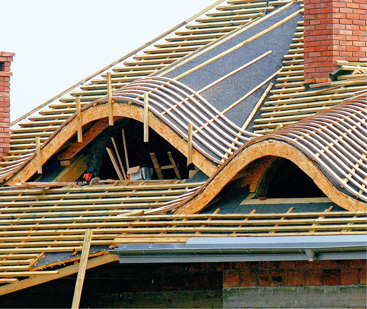 Konstrukcja dachu z lukarnami w trakcie budowy, widoczne drewniane łaty, papa i fragment komina z czerwonej cegły.