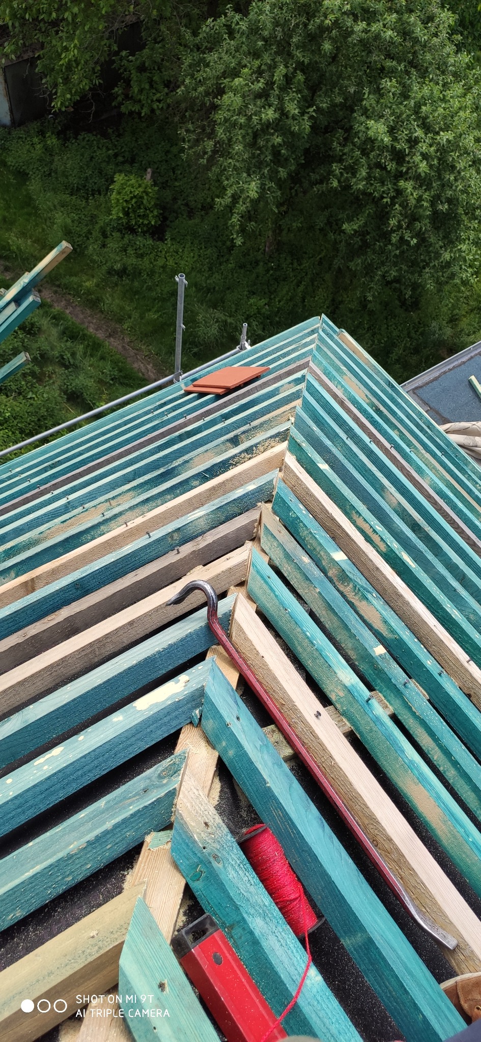 Konstrukcja więźby dachowej z pomalowanymi na niebiesko łatami, z widocznymi narzędziami: łomem i sznurkiem murarskim, częściowo pokryta dachówkami.