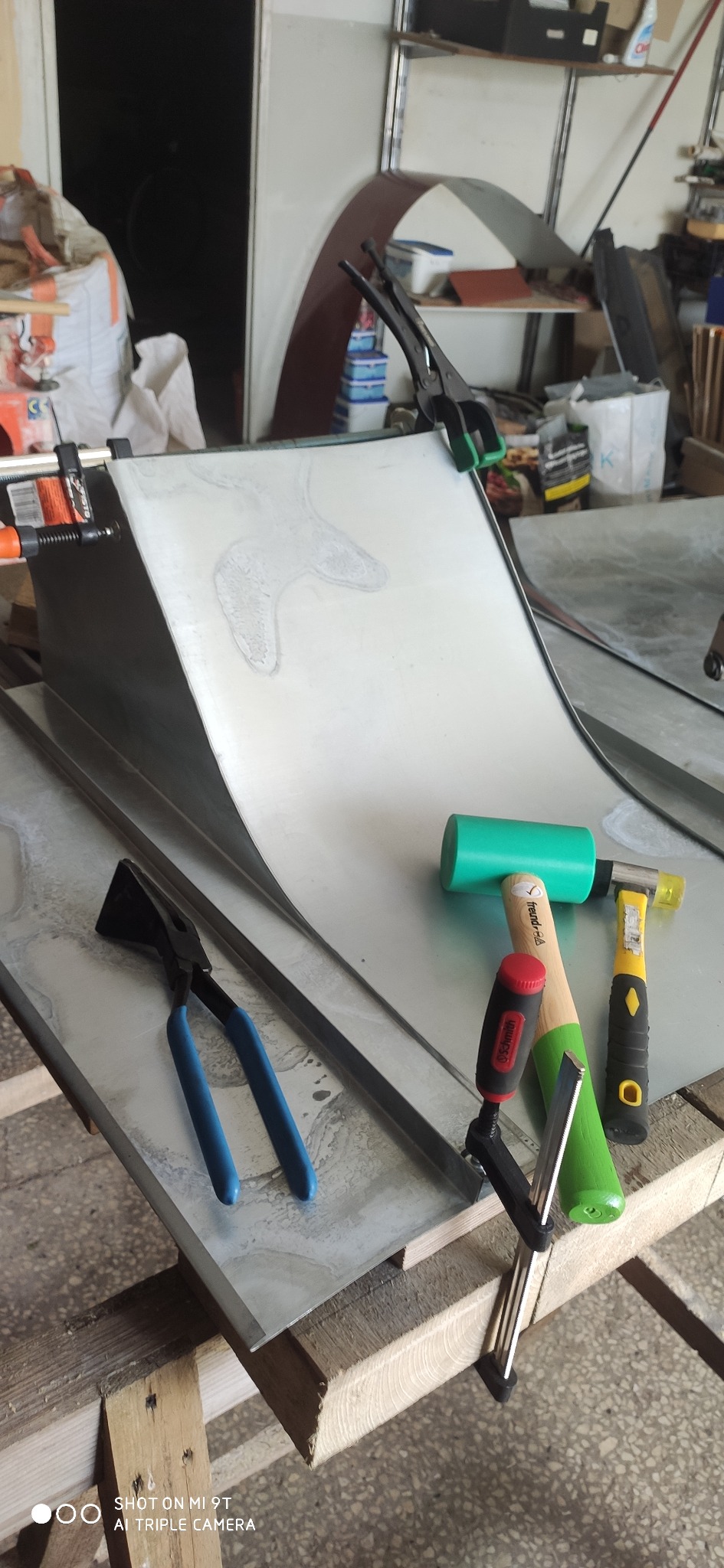 Blacha mocowana klamrami na drewnianym stole warsztatowym, obok leżą szczypce i młotki z gumowymi i plastikowymi końcówkami.