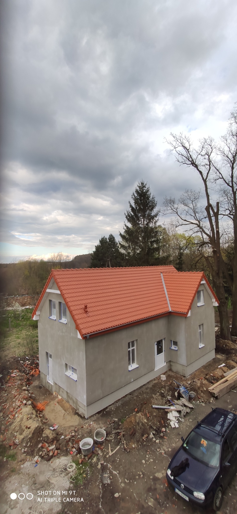 Surowy, dwukondygnacyjny dom z nowym, pomarańczowym pokryciem dachowym z dachówki ceramicznej, świeżo otynkowany, z widocznymi śladami prac budowlanych wokół budynku i samochodem osobowym...