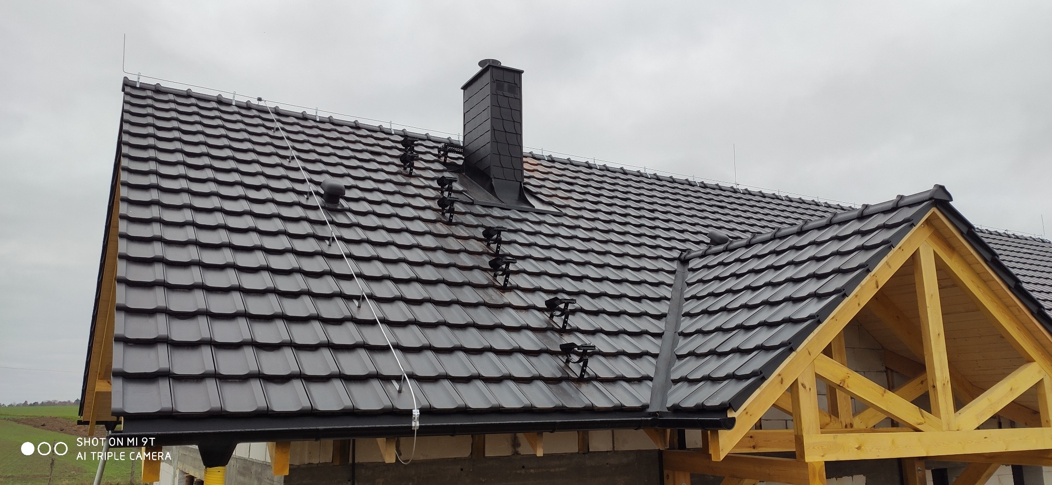 Nowy dach z czarnej dachówki ceramicznej z kominem i systemem odgromowym, widoczne drewniane elementy konstrukcji dachu w surowym stanie.