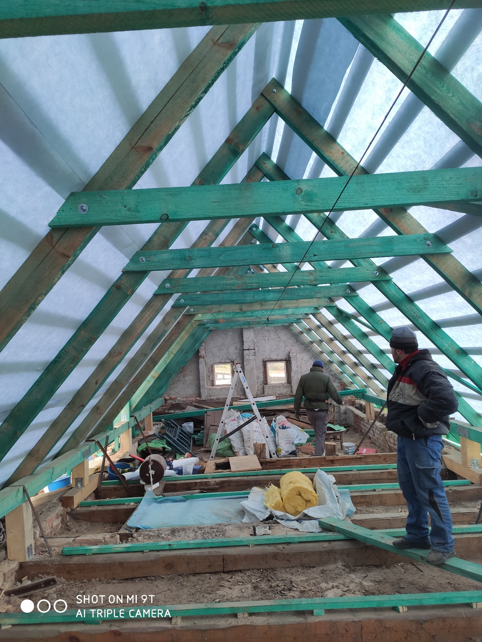 Wnętrze remontowanego poddasza z widoczną konstrukcją więźby dachowej, ociepleniem z wełny mineralnej i dwoma pracownikami w trakcie prac.