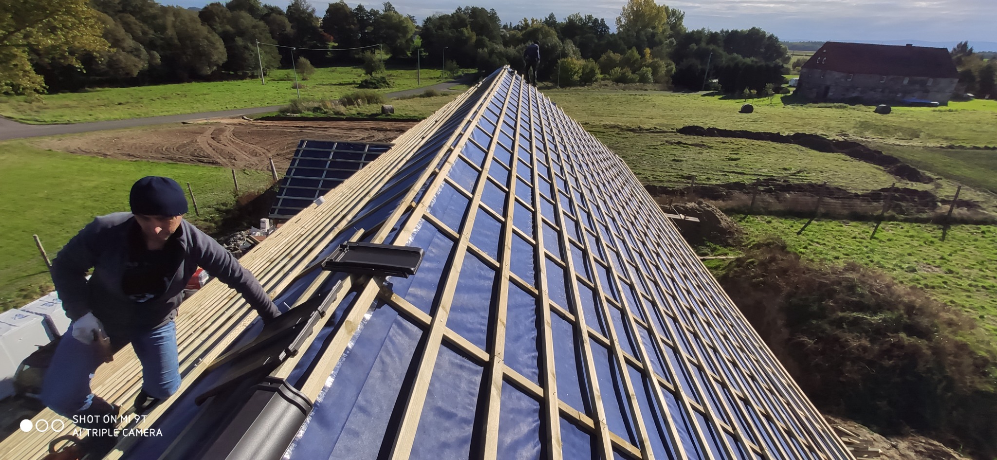 Montaż dachówki na drewnianej konstrukcji dachu pokrytego folią w Zgorzelcu, widok z góry na pracownika przy pracy i rozległy krajobraz w tle.