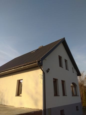 Jasna elewacja domu jednorodzinnego z ciemną, skośną połacią dachu i systemem rynnowym w kolorze grafitowym, oświetlona światłem słonecznym.