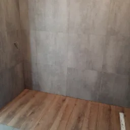 Surowa łazienka w trakcie wykańczania: szare, kwadratowe płytki ścienne i podłoga z paneli imitujących drewno, widoczny odpływ liniowy.