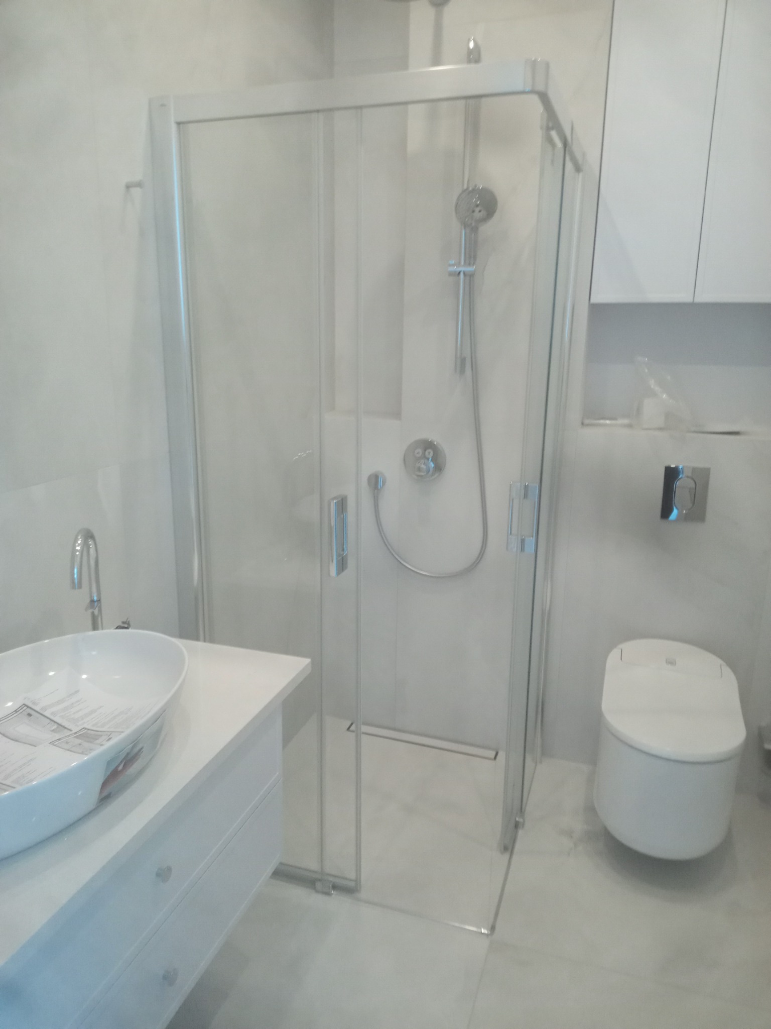 Nowoczesna, jasna łazienka z kabiną prysznicową ze szklanymi drzwiami, umywalką nablatową na białej szafce z szufladami i wiszącą toaletą.