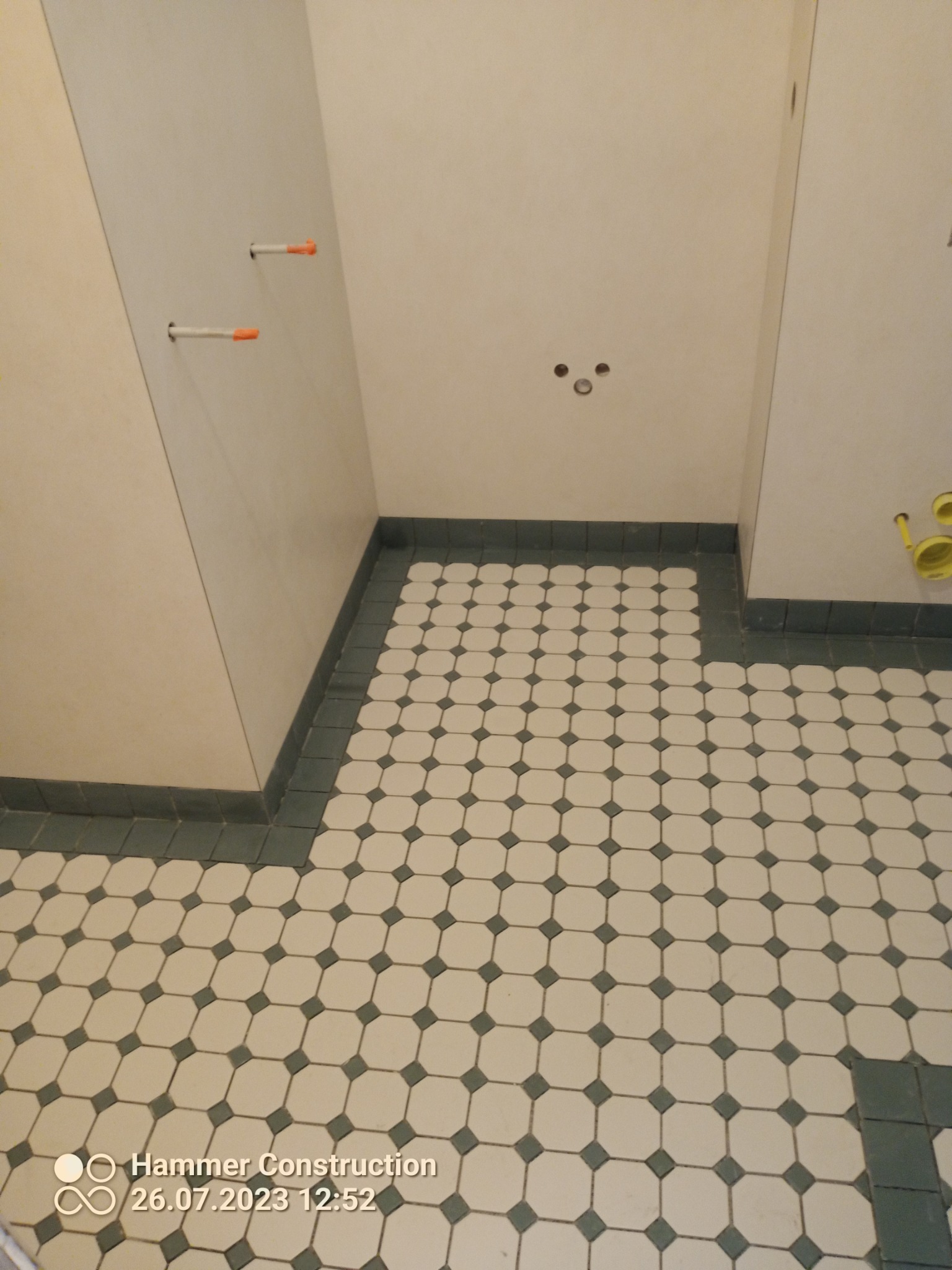 Ułożona podłoga z płytek w łazience, widoczne otwory w ścianie na instalacje, w tle fragment instalacji kanalizacyjnej.