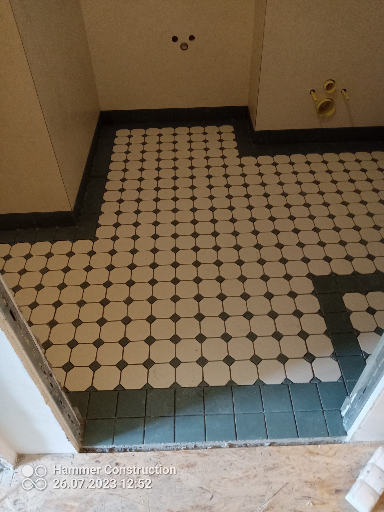 Ułożona podłoga z płytek ceramicznych w stylu retro, biało-czarne ośmiokąty z czarną obwódką, widoczne fragmenty ścian i instalacji hydraulicznej w łazience przed montażem armatury.