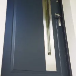 Grafitowe drzwi zewnętrzne z pionowym, wąskim przeszkleniem w stalowej ramie, widoczne ślady montażu u dołu.