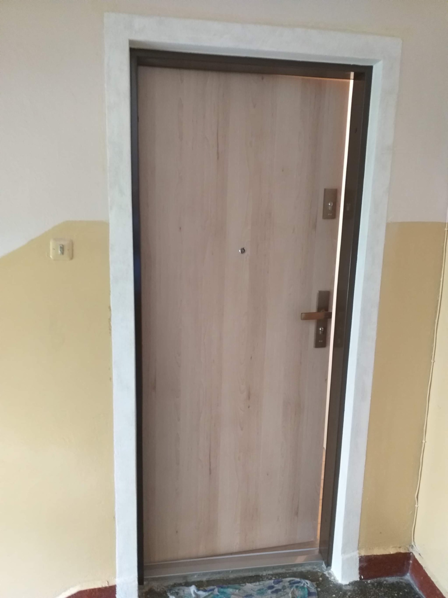 Drewnopodobne drzwi wejściowe z wizjerem i zamkiem wielopunktowym, osadzone w ościeżnicy z białym obramowaniem, widoczne od strony korytarza z żółtymi ścianami i fragmentem podłogi.