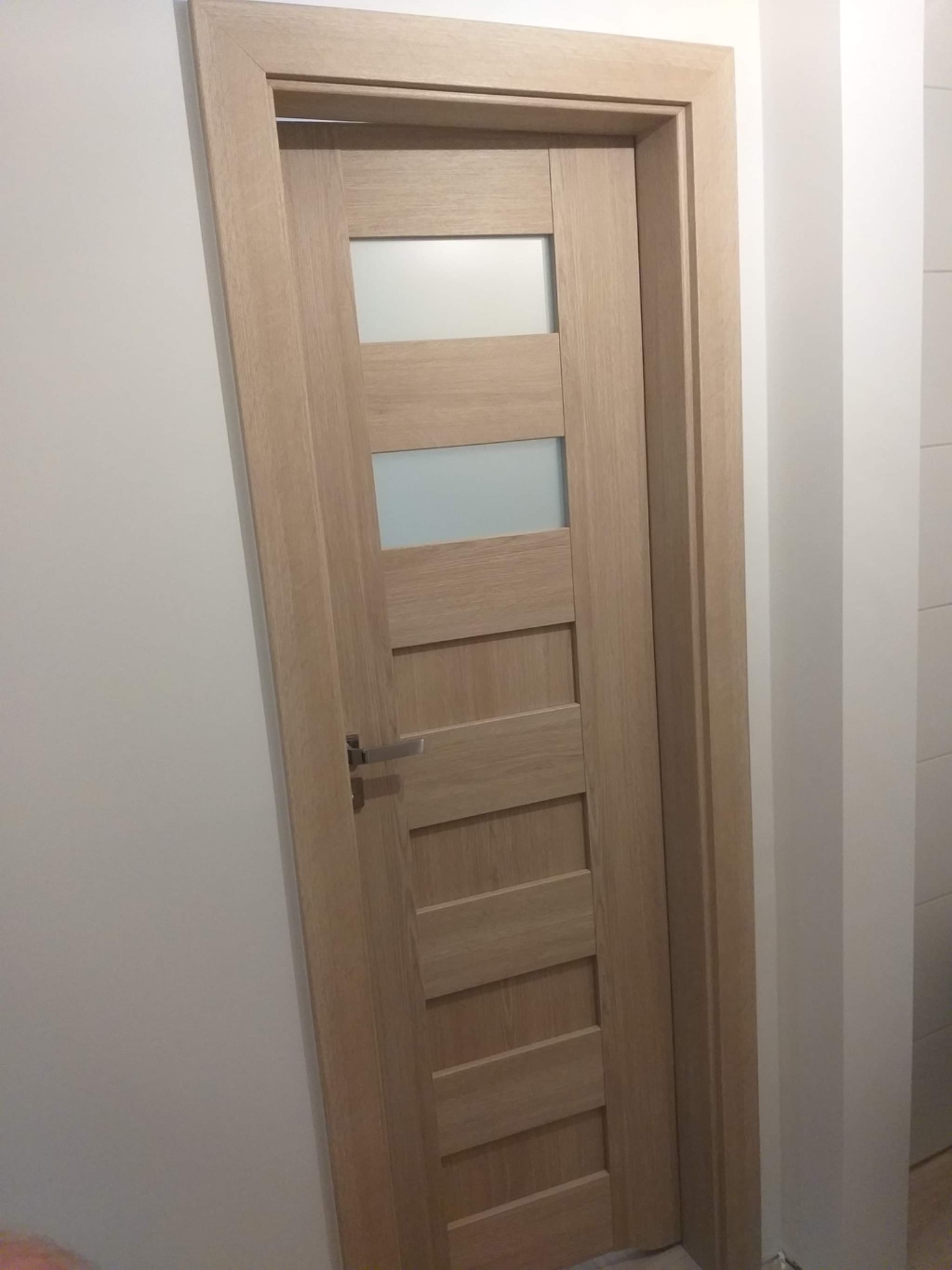 Zamknięte, drewniane drzwi wewnętrzne z czterema poziomymi, matowymi przeszkleniami, widoczne od strony klamki w minimalistycznym wnętrzu z białymi ścianami.