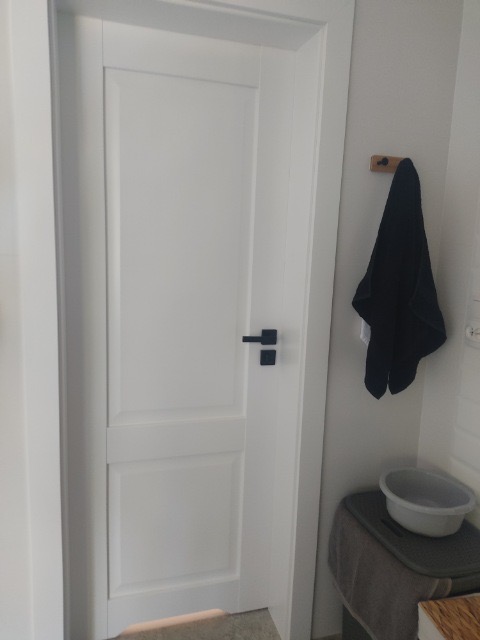 Białe drzwi wewnętrzne z czarną klamką w minimalistycznej łazience, widoczny wieszak z ręcznikiem i miska.