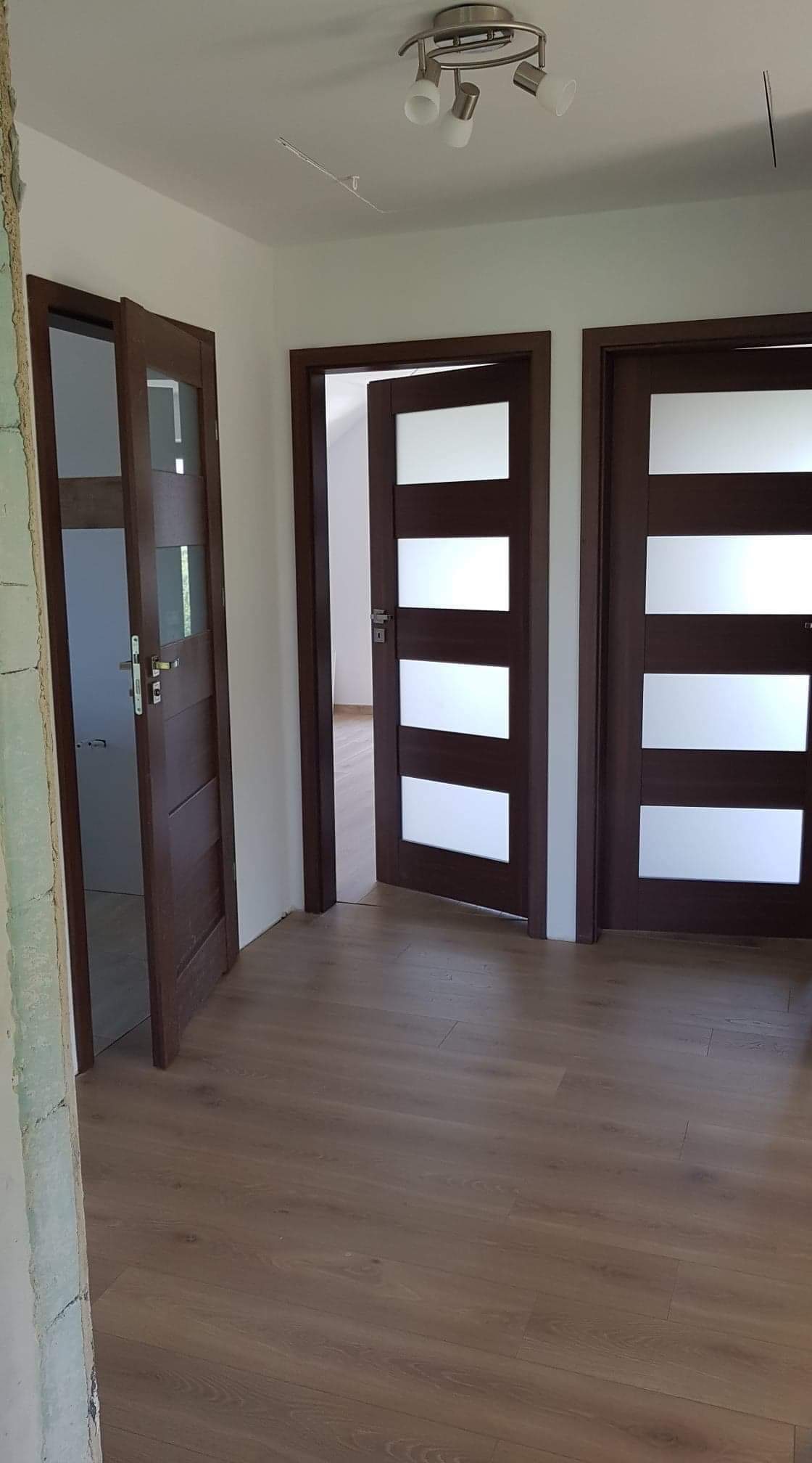 Trzy brązowe drzwi wewnętrzne z poziomymi, matowymi szybami, zamontowane na podłodze z paneli; jeden z otworów drzwiowych w trakcie wykończenia.