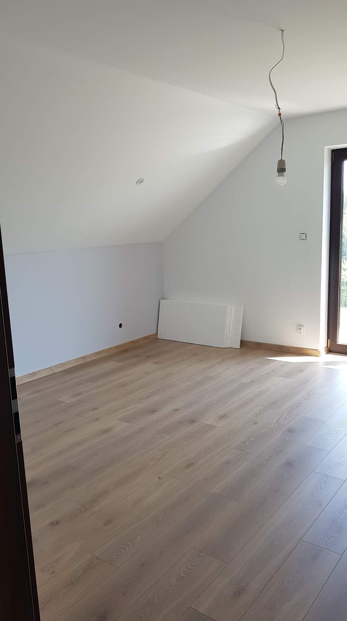Poddasze z ułożonymi panelami podłogowymi w odcieniach beżu, widoczne białe ściany, sufit ze skosem, okno z ciemną ramą i wisząca żarówka bez klosza. Przy ścianie leżą białe płyty.