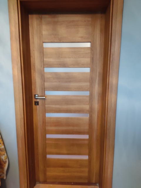 Drewniane drzwi wewnętrzne z poziomymi, przezroczystymi wstawkami, widoczna klamka ze stali nierdzewnej, ościeżnica z litego drewna.