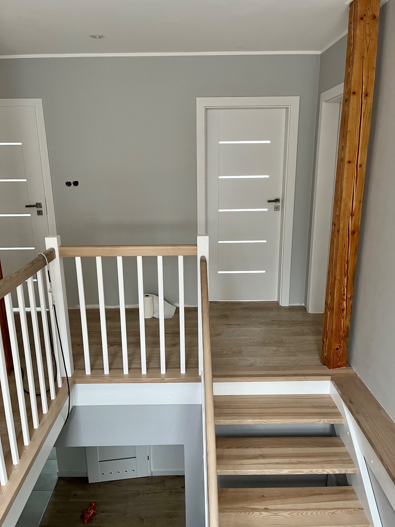Wnętrze domu po remoncie: widok na schody z drewnianymi stopniami, białą balustradą i dwoje białych drzwi wewnętrznych z poziomymi przeszkleniami, drewniany filar przy ścianie.